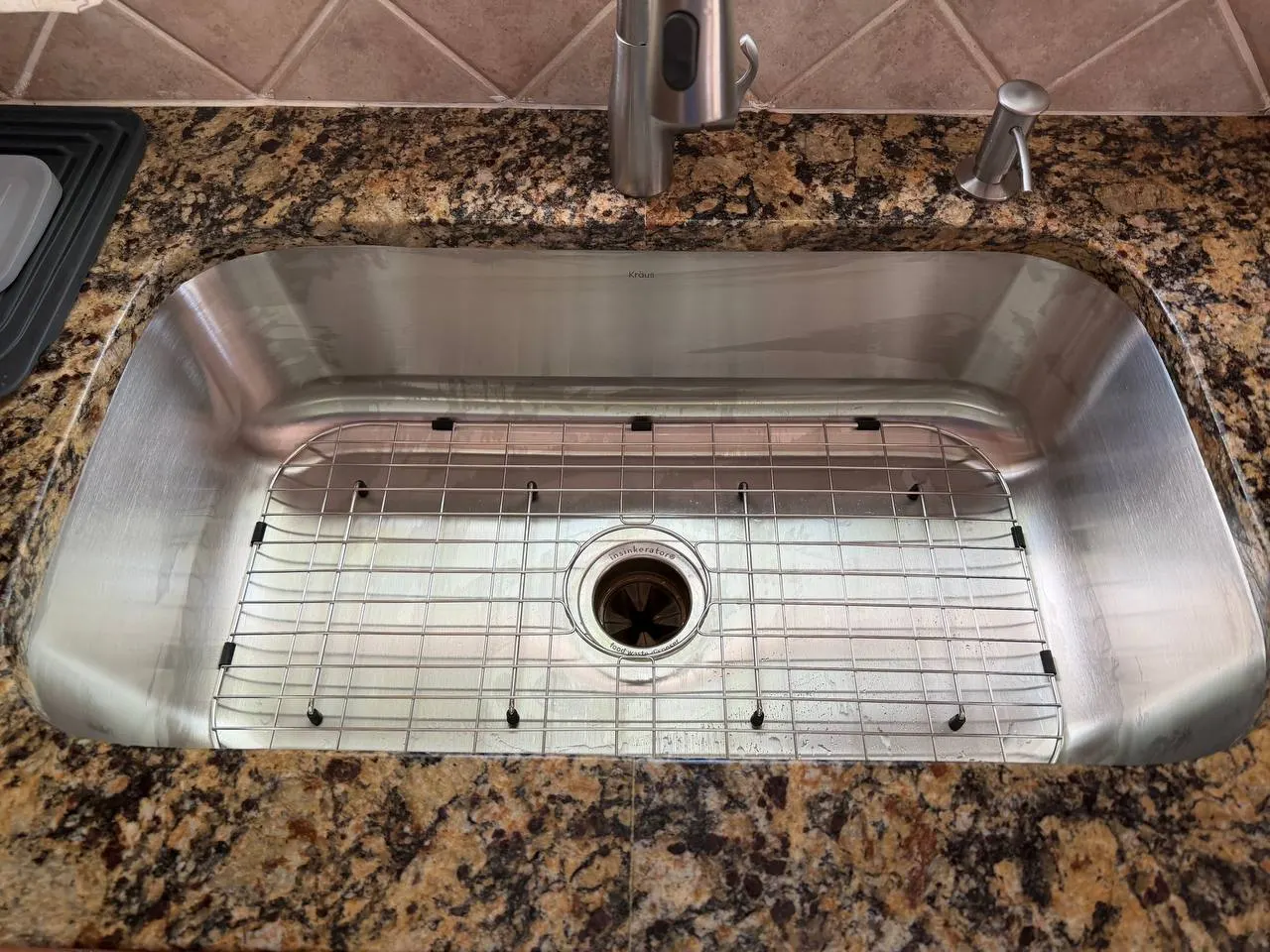 New single-bowl stainless steel undermount sink with a bottom grid and centre drain, set under the existing granite countertop
